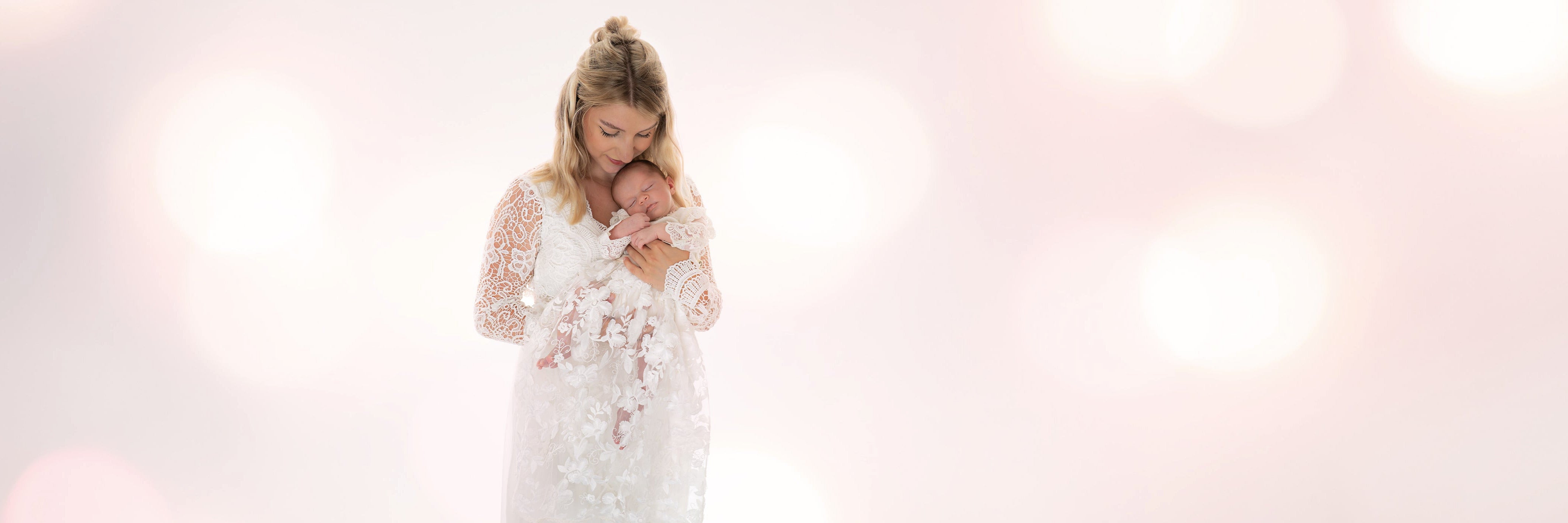 Mama hält ein neugeborenes Mädchen in einem CUCOONU Taufgewand, bestehend aus festlichem Babybody in Offwhite und langem Rock mit 3D-Stickerei