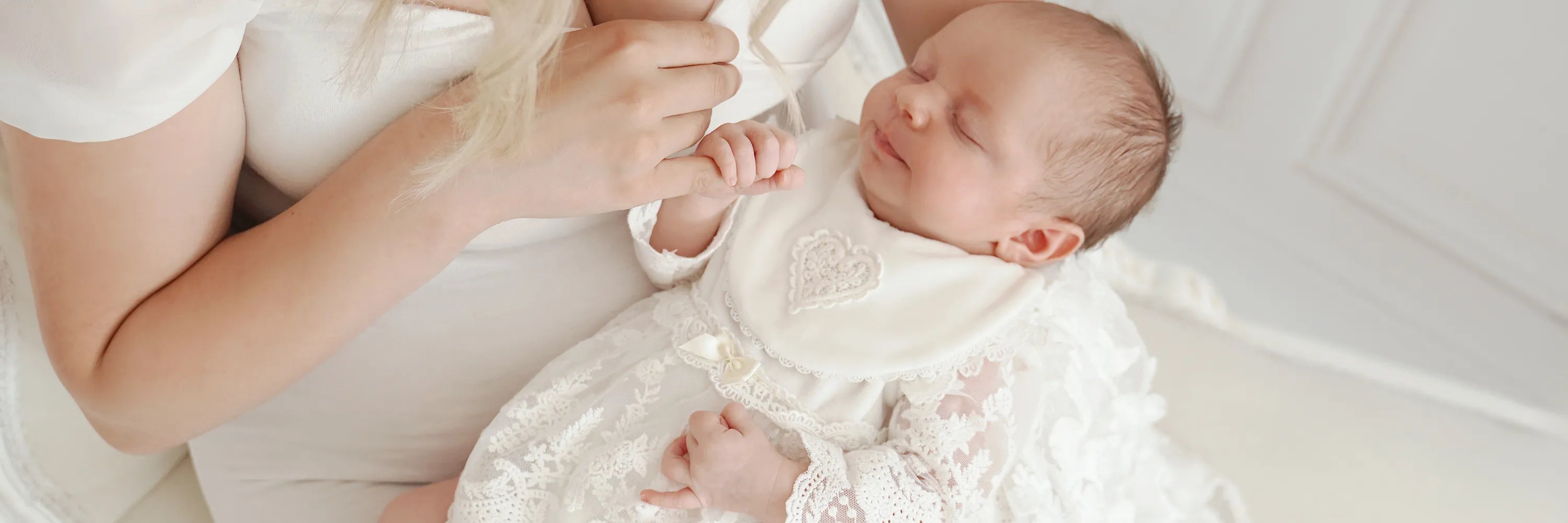Neugeborenes Mädchen trägt festliches CUCOONU Taufkleid und hält den Finger der Mutter.